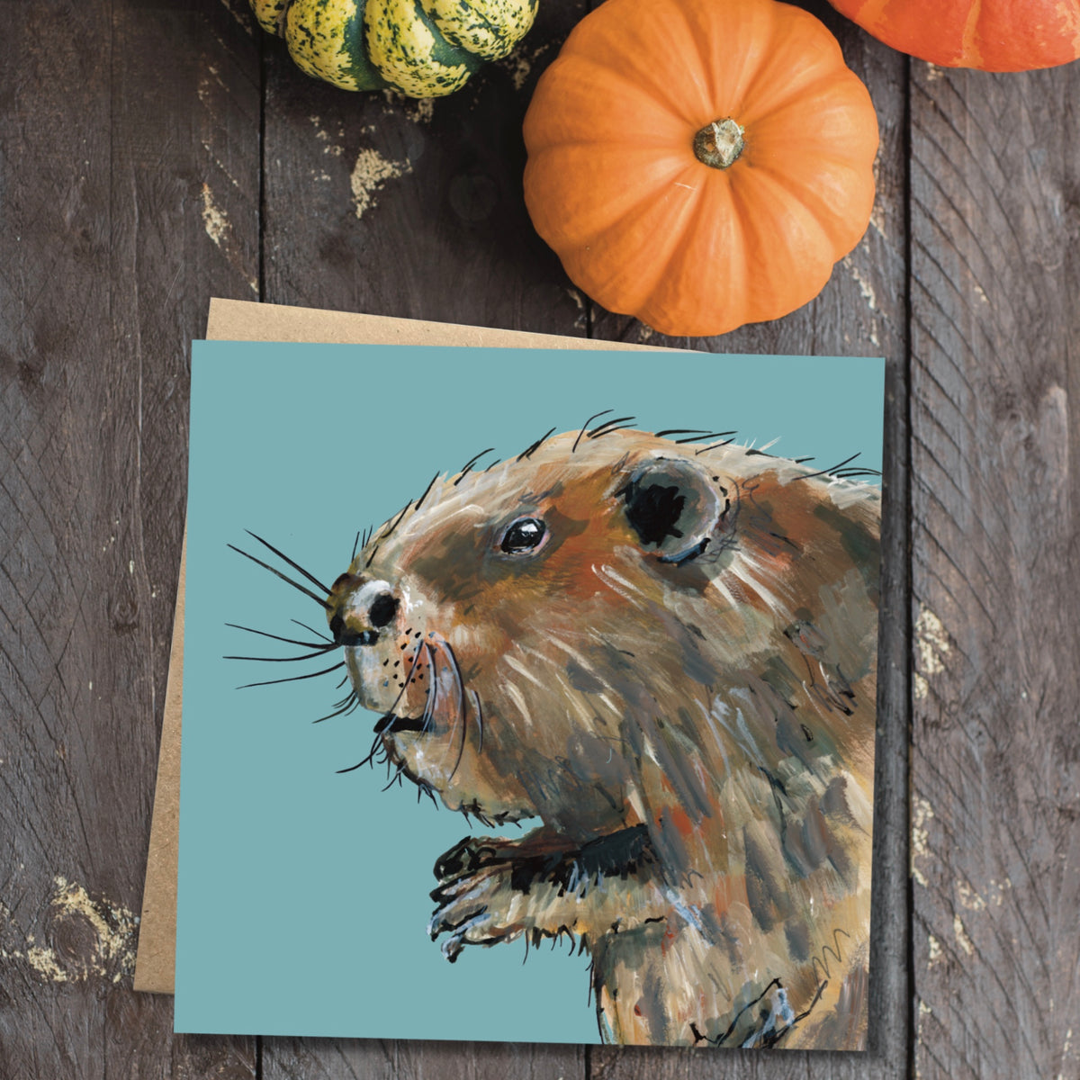 A square wildlife greetings card showing a painted brown beaver on a blue background. Along side is the cards recycled brown kraft envelope.